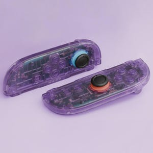 Puede incluir: Dos mandos de juego morados transparentes con componentes internos visibles. Cada mando tiene un joystick negro y botones de colores. Un joystick tiene un anillo azul, el otro un anillo naranja.