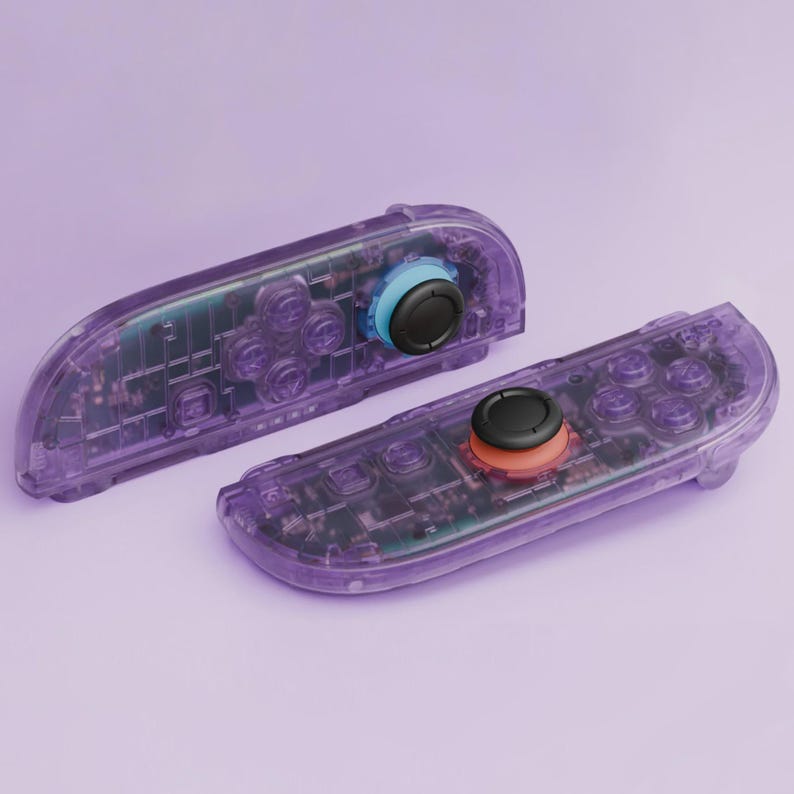 Puede incluir: Dos mandos de juego morados transparentes con componentes internos visibles. Cada mando tiene un joystick negro y botones de colores. Un joystick tiene un anillo azul, el otro un anillo naranja.