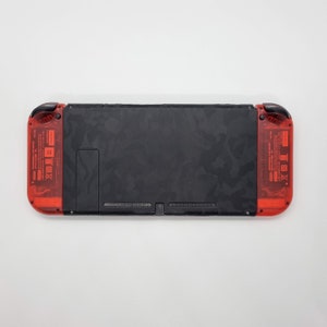 Puede incluir: Una consola Nintendo Switch negra con una piel de camuflaje. La consola tiene paneles laterales transl&uacute;cidos rojos con la marca Nintendo y un peque&ntilde;o logotipo negro de Nintendo en la parte delantera.