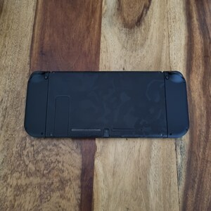 Puede incluir: Consola Nintendo Switch negra, vista desde la parte trasera, sobre una superficie de madera. La consola tiene forma rectangular con bordes redondeados y mandos acoplados. La parte trasera tiene un soporte y varios puertos. Mide unos 24 cm de largo.