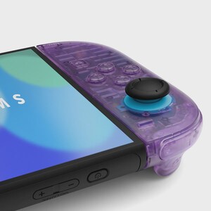 Puede incluir: Primer plano de un mando de juego transparente p&uacute;rpura con un joystick negro y detalles en azul. El mando est&aacute; conectado a un dispositivo negro con una pantalla que muestra un degradado azul y verde. La palabra "MS" es visible en la pantalla.