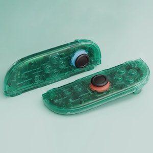 Könnte beinhalten: Zwei transparente grüne Gamecontroller mit sichtbaren internen Komponenten. Einer hat einen blauen Ring um den Joystick, der andere einen roten Ring. Beide haben schwarze Joysticks und verschiedene Tasten.