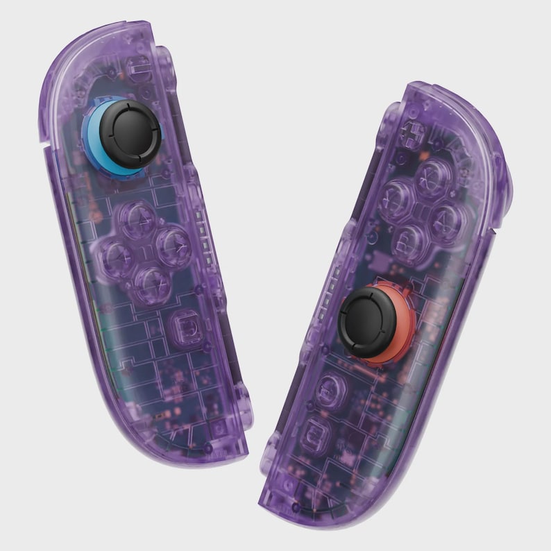 Puede incluir: Dos mandos de juego morados transparentes con componentes internos visibles. Un mando tiene un joystick negro con base azul, el otro tiene un joystick negro con base naranja. Los mandos tienen m&uacute;ltiples botones y pads direccionales.