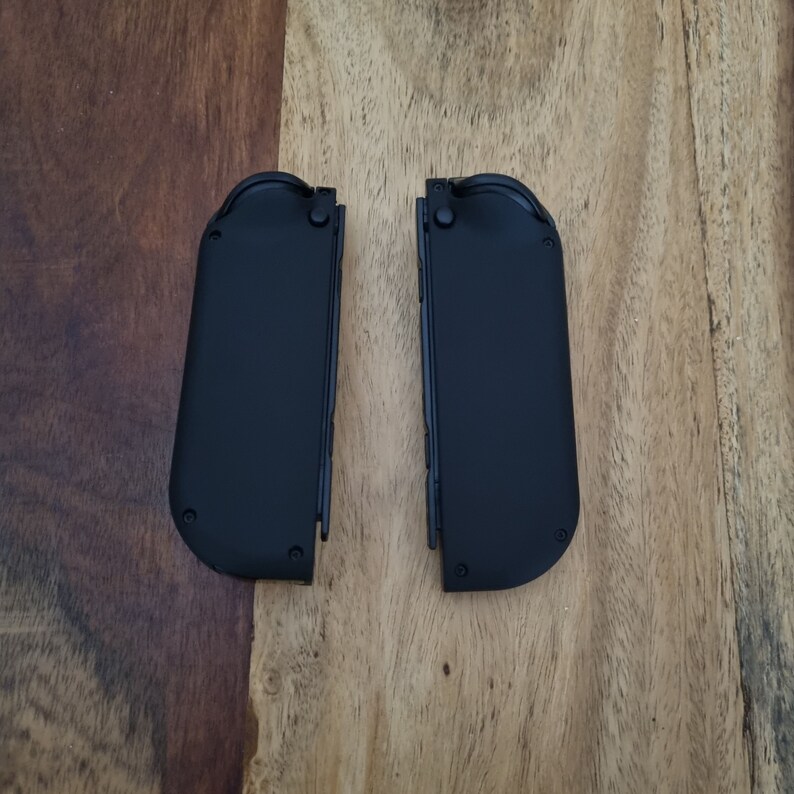 Puede incluir: Dos mandos de juego negros, probablemente para una consola, se muestran sobre un fondo de madera. Los mandos son rectangulares con bordes redondeados y peque&ntilde;os botones. Los mandos est&aacute;n uno al lado del otro.