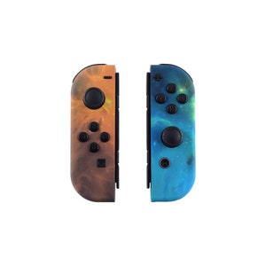 Puede incluir: Dos mandos Joy-Con para Nintendo Switch con diseños de temática galáctica. Un mando es naranja y marrón con un patrón de nebulosa, el otro es azul y verde con un patrón de nebulosa.