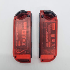 Puede incluir: Dos carcasas de consola de juegos Nintendo Switch rojas con botones negros y una parte posterior de pl&aacute;stico transparente. La parte posterior de las carcasas tiene el texto "RECHARGEABLE LI-ION BATTERY" y "リチウムイオン電池" en japon&eacute;s. El texto "HAC-006" est&aacute; impreso en ambas carcasas. El texto "PWRB18D21" est&aacute; impreso en la esquina inferior derecha de cada carcasa.