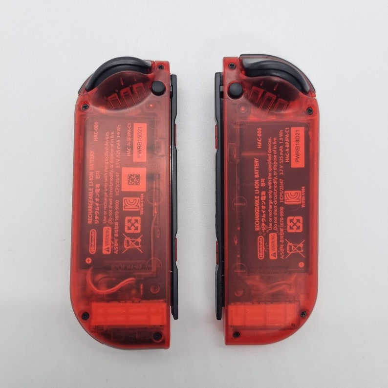 Puede incluir: Dos carcasas de consola de juegos Nintendo Switch rojas con botones negros y una parte posterior de pl&aacute;stico transparente. La parte posterior de las carcasas tiene el texto "RECHARGEABLE LI-ION BATTERY" y "リチウムイオン電池" en japon&eacute;s. El texto "HAC-006" est&aacute; impreso en ambas carcasas. El texto "PWRB18D21" est&aacute; impreso en la esquina inferior derecha de cada carcasa.