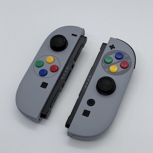 Könnte beinhalten: Ein Paar graue Videospiel-Controller mit schwarzen Analogsticks und vier bunten Tasten in Rot, Blau, Gelb und Grün. Der rechte Controller verfügt über eine Home-Taste, eine Aufnahmetaste und ein Plus-Symbol.