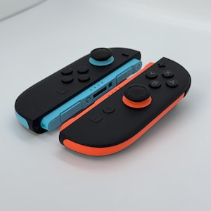 May include: Two black and colored game controllers. One controller has a black joystick and buttons with a blue base. The other controller has a black joystick and buttons with an orange base. The controllers are for a gaming console.