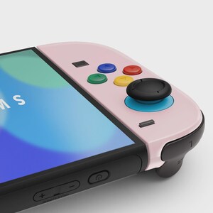 May include: A pink and black handheld gaming console with colorful buttons. The console features a screen displaying a blue and white graphic. The buttons are blue, red, green, and yellow. The joystick is black with a blue base.