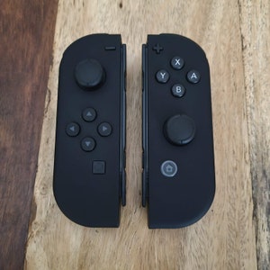 May include: Two black game controllers, each with a joystick and buttons, are displayed side-by-side. The controllers have a matte finish and are set against a wooden surface. The buttons include directional pads, A, B, X, and Y.