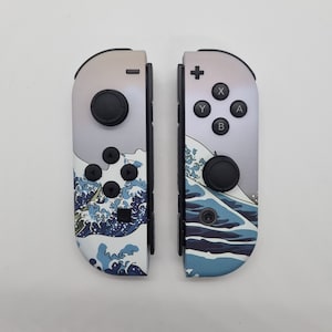 May include: Two Nintendo Switch Joy-Con controllers with a custom design. The controllers feature a wave pattern in shades of blue and white, with black buttons and joysticks. The design is inspired by Japanese art.