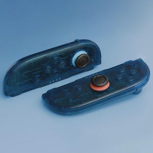 Könnte beinhalten: Zwei transparente blaue Gamecontroller mit sichtbaren internen Komponenten. Die Controller haben schwarze Joysticks mit blauen und orangefarbenen Akzenten. Die Tasten und Richtungspads sind durch den durchscheinenden Kunststoff sichtbar.