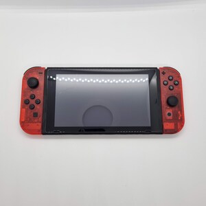 Puede incluir: Una consola Nintendo Switch roja y negra con una carcasa de pl&aacute;stico transparente. La consola tiene una pantalla negra y botones.