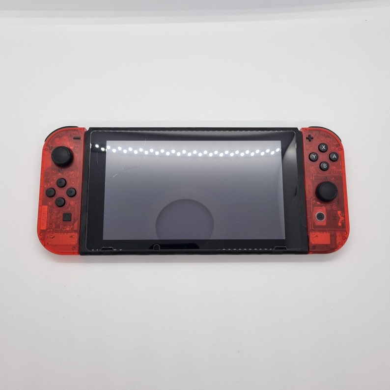 Puede incluir: Una consola Nintendo Switch roja y negra con una carcasa de pl&aacute;stico transparente. La consola tiene una pantalla negra y botones.