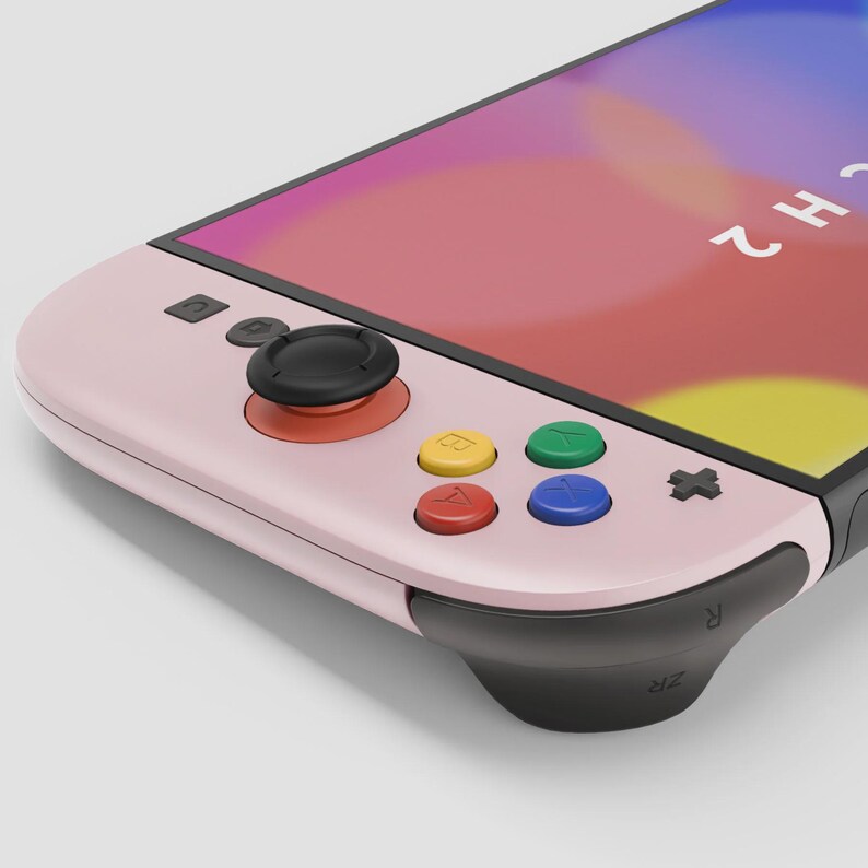 May include: A pink handheld gaming console with a screen displaying a colorful graphic. The console features a black joystick, colored buttons, and black accents. The buttons are red, yellow, green, and blue.