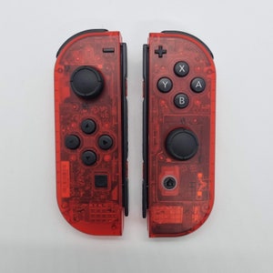 Puede incluir: Un par de mandos Nintendo Switch Joy-Con transparentes rojos. Los mandos tienen botones negros y una palanca de mando negra.