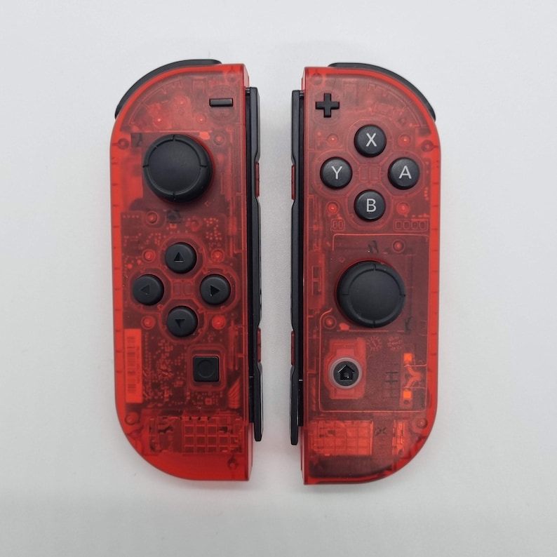Puede incluir: Un par de mandos Nintendo Switch Joy-Con transparentes rojos. Los mandos tienen botones negros y una palanca de mando negra.