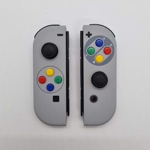 May include: A pair of gray Nintendo Switch controllers with custom face buttons. The buttons are colored red, yellow, blue, and green. The controllers have a black border and a black button on the front.
