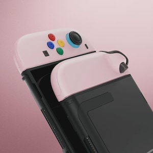 May include: A pink and black handheld gaming console with colorful buttons. The console features a pink top with red, blue, yellow, and green buttons, a black screen, and a black handle. The console is designed for gaming.