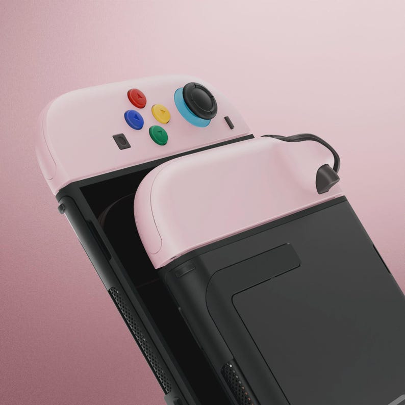 May include: A pink and black handheld gaming console with colorful buttons. The console features a pink top with red, blue, yellow, and green buttons, a black screen, and a black handle. The console is designed for gaming.