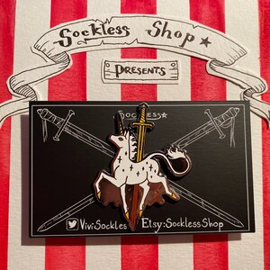 Puede incluir: Un unicornio blanco con detalles dorados es atravesado por una espada. El unicornio está de pie sobre una base marrón. El pin está sobre un fondo negro con texto blanco que dice "Sockless Shop Presents" y "ViviSockles Etsy:SocklessShop".