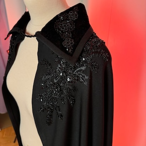 Op de afbeelding: Een zwarte cape met een fluwelen kraag en zwarte parelversieringen. De cape heeft een puntige kraag en een lang, vloeiende ontwerp.