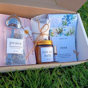 May include: A gift box filled with self-care items. Includes a tea infuser, a jar labeled "you are held", a small essential oil bottle, a floral handkerchief, and a card with the word "rest". The box is set on a bed of green grass.