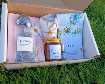 May include: A gift box filled with self-care items. Includes a tea infuser, a jar labeled "you are held", a small essential oil bottle, a floral handkerchief, and a card with the word "rest". The box is set on a bed of green grass.