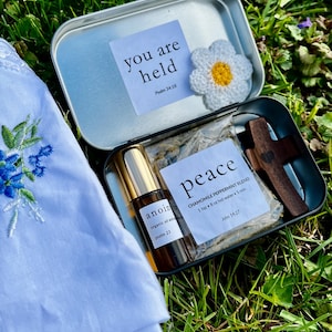 May include: A small, open metal tin contains a bottle of anointing oil, a wooden cross, and a packet labeled "peace." A crocheted daisy and a card with the text "you are held" are also inside. A white cloth with blue floral embroidery is in the foreground.