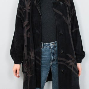 Puede incluir: Un abrigo negro con un dise&ntilde;o de hojas de bamb&uacute; marr&oacute;n oscuro. El abrigo tiene una botonadura frontal, mangas largas y un cuello. La persona lleva un cuello alto gris y vaqueros azules.