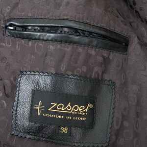 Puede incluir: Primer plano del interior de una chaqueta de cuero negra. Un bolsillo negro con cremallera es visible en la parte superior. Una etiqueta negra con texto dorado dice "zaspe design COUTURE IN LEDER" y "38". El forro tiene un patr&oacute;n oscuro.