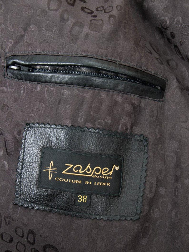 Puede incluir: Primer plano del interior de una chaqueta de cuero negra. Un bolsillo negro con cremallera es visible en la parte superior. Una etiqueta negra con texto dorado dice "zaspe design COUTURE IN LEDER" y "38". El forro tiene un patr&oacute;n oscuro.