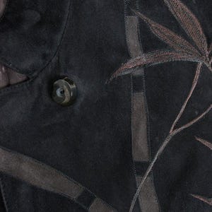 Puede incluir: Primer plano de una chaqueta de ante negra con dise&ntilde;o de hojas bordadas en marr&oacute;n. La chaqueta presenta un bot&oacute;n grande y redondo y detalles de ante marr&oacute;n. El dise&ntilde;o incluye l&iacute;neas marrones cosidas y un patr&oacute;n de hojas.