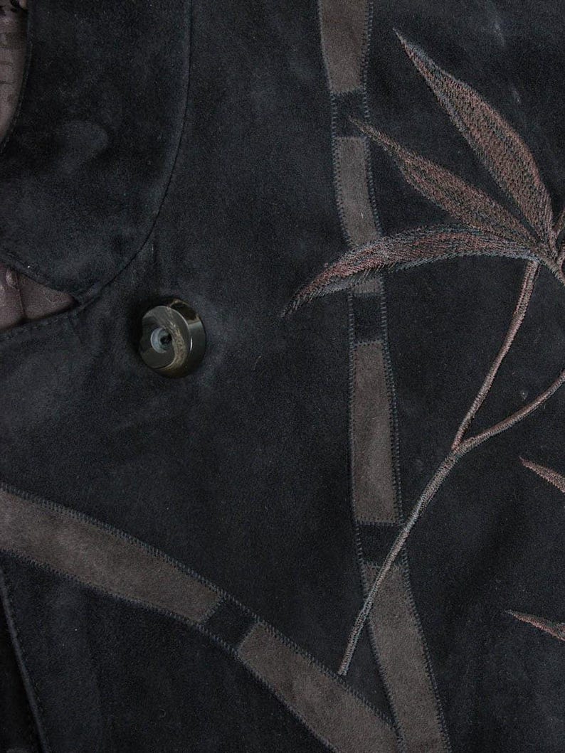 Puede incluir: Primer plano de una chaqueta de ante negra con dise&ntilde;o de hojas bordadas en marr&oacute;n. La chaqueta presenta un bot&oacute;n grande y redondo y detalles de ante marr&oacute;n. El dise&ntilde;o incluye l&iacute;neas marrones cosidas y un patr&oacute;n de hojas.
