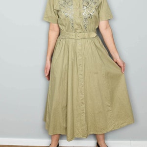 Könnte beinhalten: Ein hell olivgrünes Kleid mit kurzen Ärmeln, Knopfleiste vorne und einem Gürtel in der Taille. Das Kleid hat gestickte florale Details auf dem Oberteil und einen ausgestellten Rock. Das Model trägt schwarze Plateausandalen.