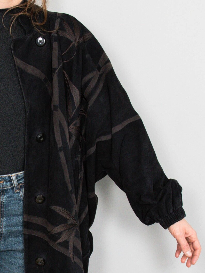 Puede incluir: Abrigo negro con botones y un estampado de hojas de bamb&uacute; marr&oacute;n. El abrigo tiene un corte holgado con pu&ntilde;o fruncido. El abrigo se lleva sobre una camiseta gris oscuro y vaqueros azules.
