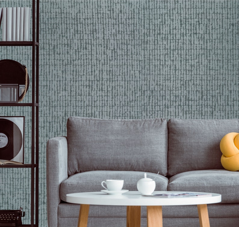 May include: A gray couch with a yellow pillow sits in front of a wall with a blue and white patterned wallpaper. A small round table with a white surface sits in front of the couch. There are two white teacups and a white sugar bowl on the table.