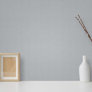 Pu&ograve; includere: Un vaso in ceramica bianca con una superficie testurizzata contiene un bouquet di rami secchi con piccoli boccioli bianchi. Una bottiglia in ceramica bianca e una cornice in legno sono su una superficie bianca davanti a un muro grigio testurizzato.