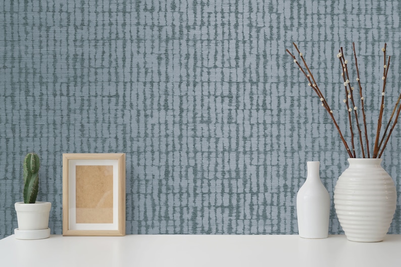 May include: A blue and white patterned wallpaper with a subtle texture. The wallpaper is displayed in a room with a picture frame, a cactus in a white pot, and a vase with branches.