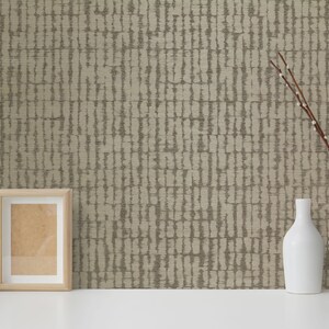 May include: A beige wallpaper with a vertical pattern of brown lines. A white picture frame, a cactus in a white pot, a white vase with branches, and a white bottle are on a white surface in front of the wallpaper.