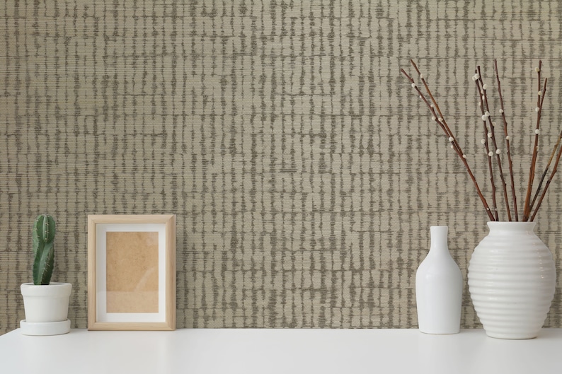 May include: A beige wallpaper with a vertical pattern of brown lines. A white picture frame, a cactus in a white pot, a white vase with branches, and a white bottle are on a white surface in front of the wallpaper.