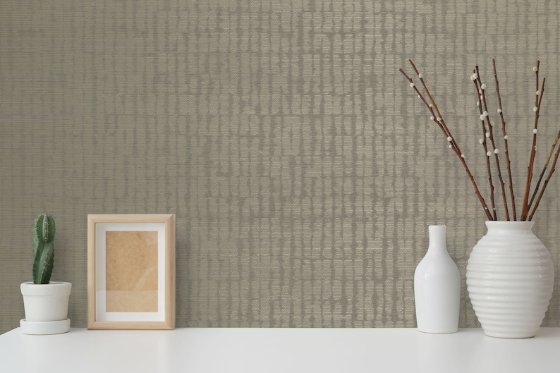 May include: A beige textured wallpaper with a subtle pattern. The wallpaper is displayed on a white surface with a picture frame, a cactus in a white pot, and a vase with branches and two white ceramic vases.