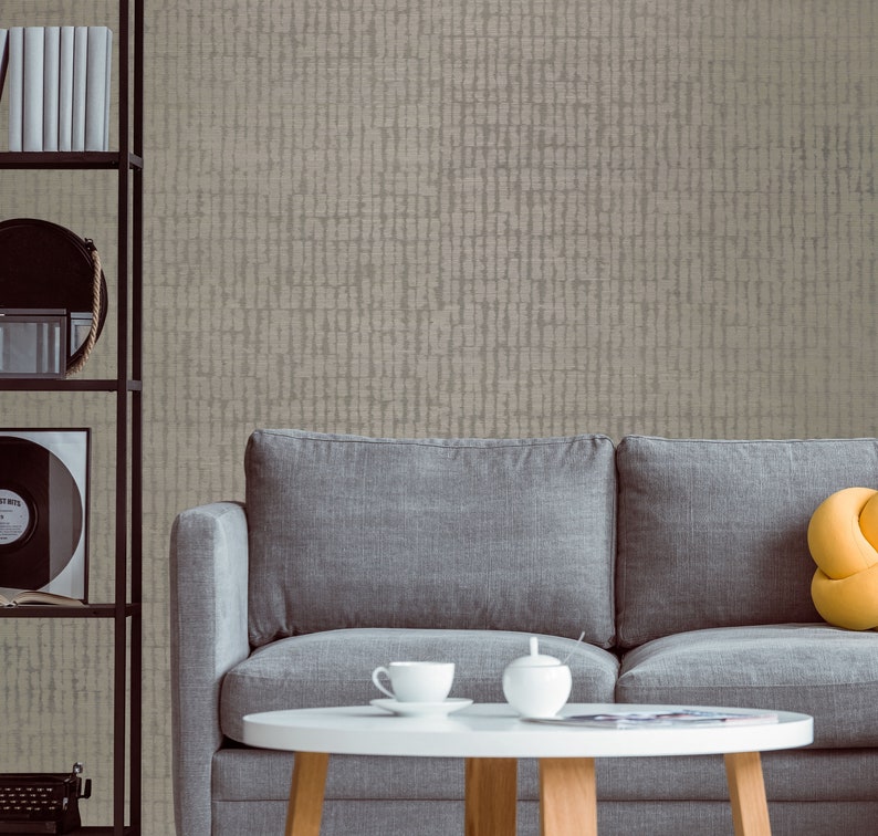 May include: A gray sofa with a yellow pillow sits in front of a wall with a textured beige wallpaper. A white coffee table with a cup and saucer and a sugar bowl sits in front of the sofa.