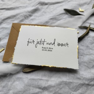 May include: A white card with gold edges and the text "f&uuml;r jetzt und immer" in black calligraphy. Below the text is "Kaja & Alicia" and "25.05.2024" in smaller black text.