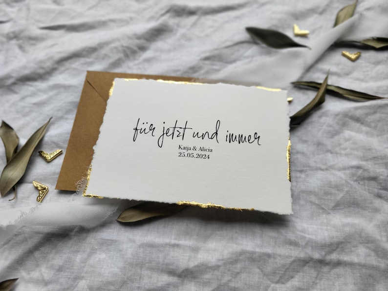 May include: A white card with gold edges and the text "f&uuml;r jetzt und immer" in black calligraphy. Below the text is "Kaja & Alicia" and "25.05.2024" in smaller black text.