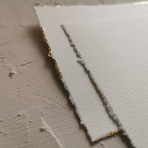 May include: Two pieces of white paper with gold edges. The paper has a textured surface and is slightly torn.