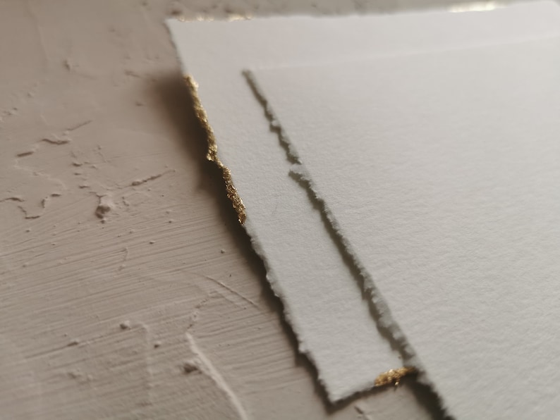 May include: Two pieces of white paper with gold edges. The paper has a textured surface and is slightly torn.