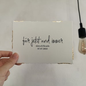 May include: A white card with gold edges and the text "f&uuml;r jetzt und immer" in black cursive writing. Below the text is "Aileen & Ricardo" and "07.07.2023" in black print.