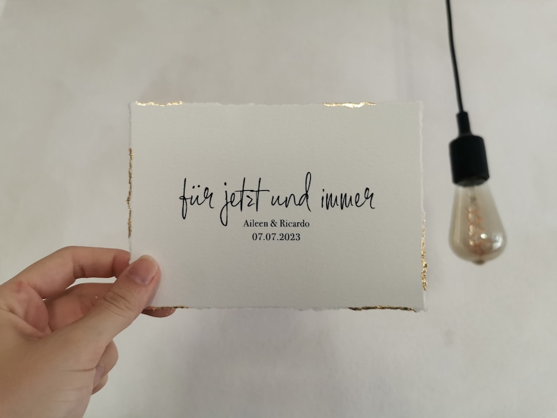 May include: A white card with gold edges and the text "f&uuml;r jetzt und immer" in black cursive writing. Below the text is "Aileen & Ricardo" and "07.07.2023" in black print.